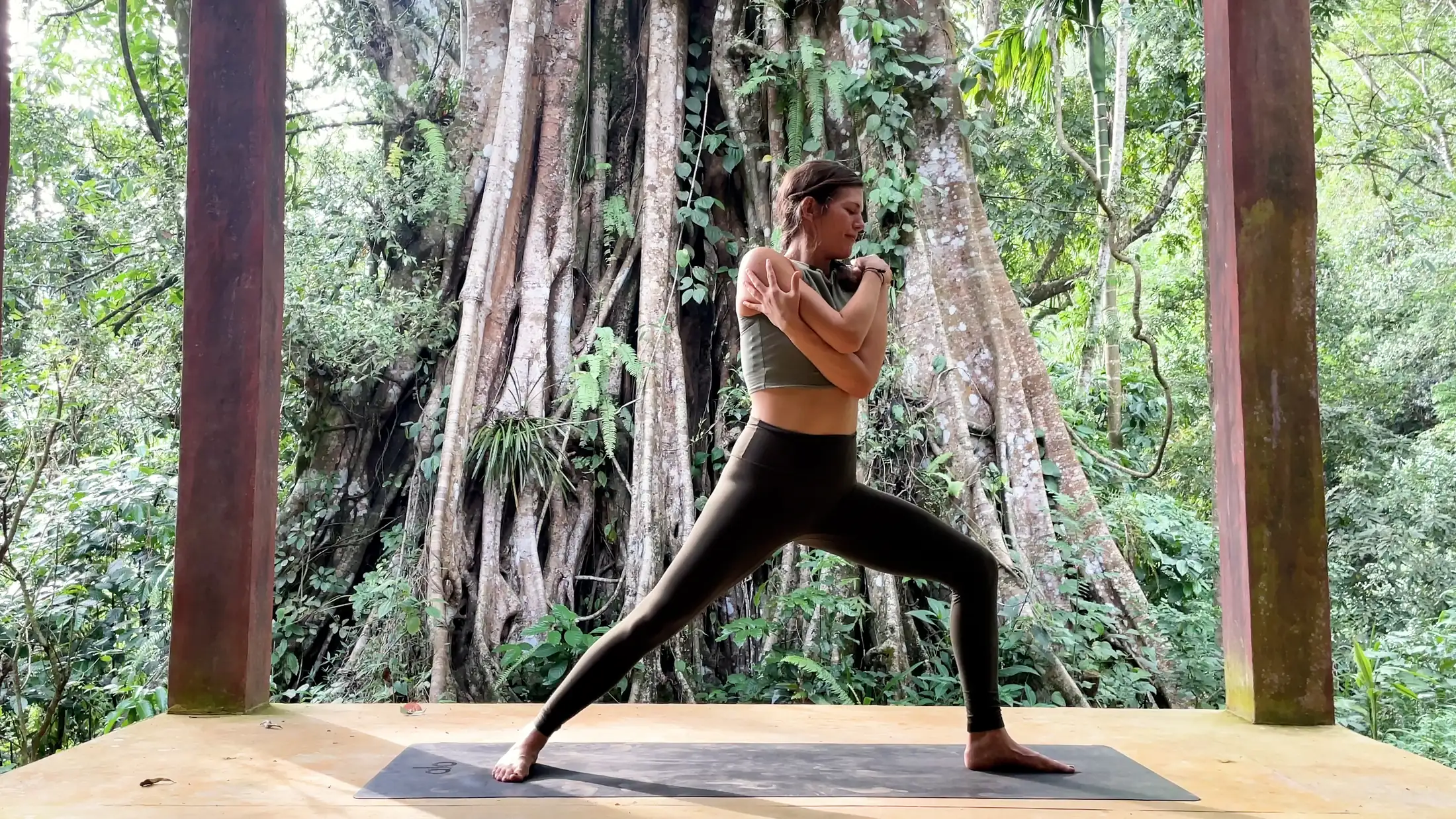 Elena Orth in a standing lunge on a wooden deck before a large banyan tree at Senses Sri Lanka